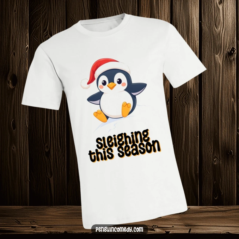 Funny holiday t-shirt showing a gleeful penguin in a Santa hat joyfully sledding down a snow-covered slope.